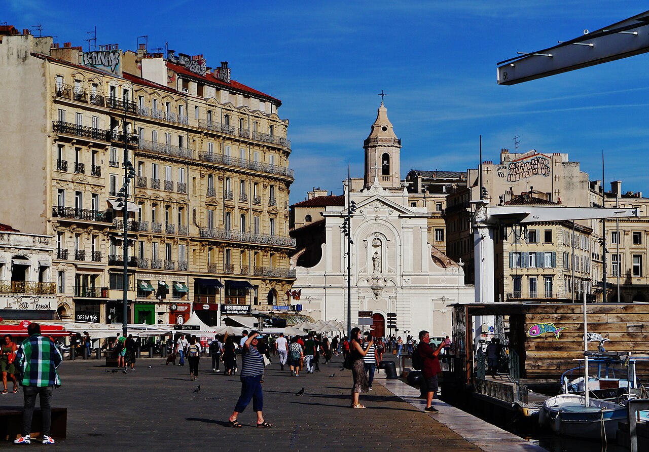 Esplanade animée du Quai du Port à Marseille : façades haussmanniennes et commerces, l'église Saint-Ferréol-les-Augustins et une barque du Vieux-Port
