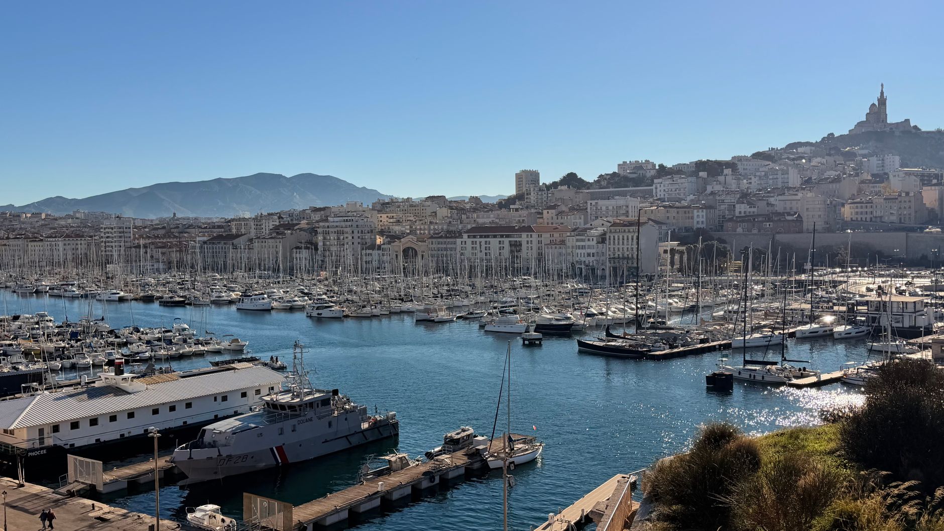 Vue dégagée du Vieux-Port de Marseille sous un ciel bleu, voiliers amarrés et Notre-Dame de la Garde sur la colline